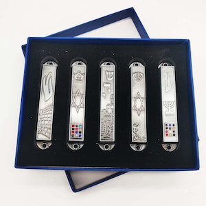 May include: A set of five silver mezuzah cases with different designs, including a menorah, a Star of David, a crown, and a Hebrew inscription. Each case has a small row of colorful rhinestones.
