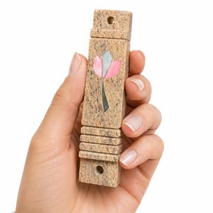 May include: A rectangular tan stone mezuzah case featuring a speckled texture and a central inlay of a stylized flower with pink and grey petals. The case has horizontal grooved detailing near the bottom and two circular holes for mounting.