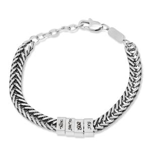 Könnte beinhalten: Silberfarbenes Armband mit geflochtenem Kettenmuster. Das Armband hat vier kleine, rechteckige Perlen mit graviertem Text. Der Verschluss und die verstellbare Kette sind ebenfalls silberfarben. Auf weißem Hintergrund.