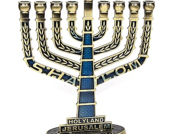 May include: A gold menorah with blue and black enamel detailing. The menorah has seven branches and is decorated with the Hebrew word "Shalom" and a design of wheat stalks. The base of the menorah has the words "Holyland Jerusalem" inscribed on it.