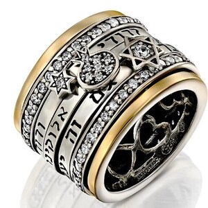 Pu&ograve; includere: Un anello in argento e oro con una fascia rotante che presenta testo ebraico, una stella di David e un piccolo cuore di cristallo trasparente. L'anello &egrave; decorato con cristalli trasparenti.