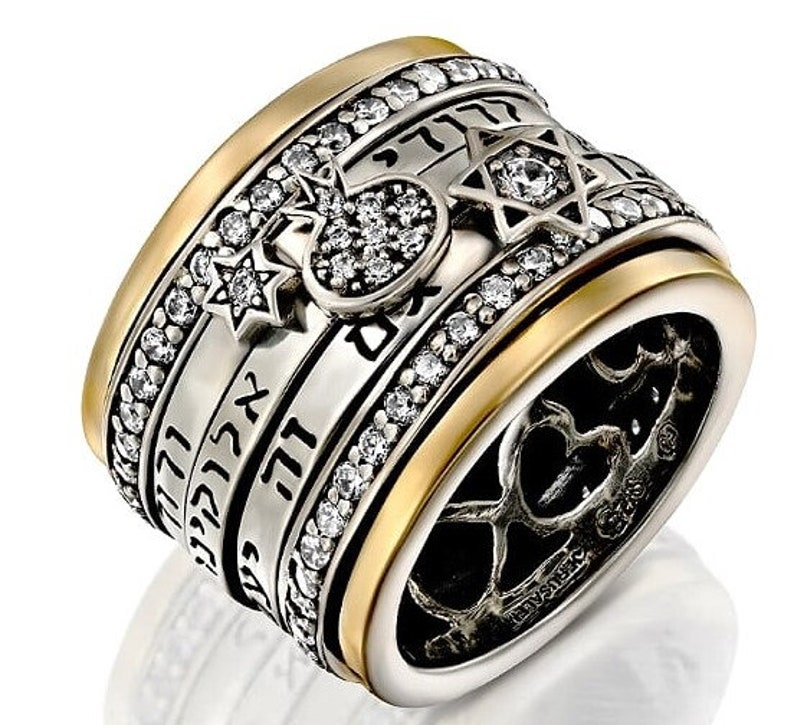 Pu&ograve; includere: Un anello in argento e oro con una fascia rotante che presenta testo ebraico, una stella di David e un piccolo cuore di cristallo trasparente. L'anello &egrave; decorato con cristalli trasparenti.