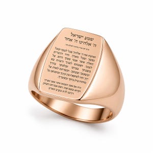 May include: A rose gold signet ring featuring a flat, polished top surface engraved with Hebrew text. The ring has a smooth, rounded band and a high-shine metallic finish. The inscription covers the entire face of the ring in black lettering.
