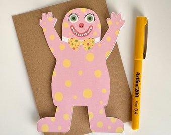 May include: A pink, cartoon-like figure with yellow polka dots, a yellow and green bow tie, and a smiling face. The figure is on a brown card with a yellow Artline 200 fine 0.4 pen next to it. The pen is yellow with black text.