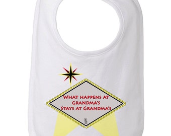 May include: White baby bib with a yellow and red sign that says "What happens at Grandma's stays at Grandma's". The sign is in the shape of a diamond and has a star above it. There are two yellow spotlights shining on the sign.