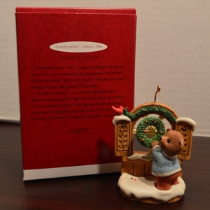 May include: A brown bear figurine wearing a blue sweater hangs a green wreath on a wooden door. The figurine is part of a Christmas ornament set. The ornament is dated 1996.
