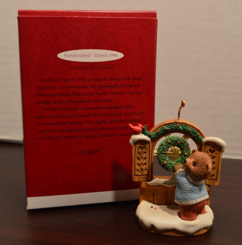 May include: A brown bear figurine wearing a blue sweater hangs a green wreath on a wooden door. The figurine is part of a Christmas ornament set. The ornament is dated 1996.