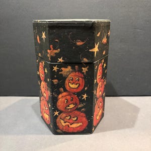 May include: A hexagonal black box featuring a stack of three orange jack-o'-lanterns on the front panel. The box is decorated with a pattern of small yellow stars and scattered autumn leaves across the black surface, including on the lid.