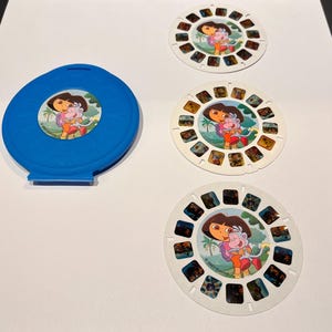 May include: A blue plastic View-Master reel storage case and three circular white View-Master reels. Each reel features a central illustration of the cartoon characters Dora and Boots hugging, with the text 2003 Viacom printed on the surface.