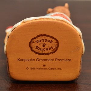 May include: A brown ceramic Christmas ornament featuring a bear wearing a red and white Santa hat and holding a red and white candy cane. The ornament is part of the "Tender Touches" collection and is a "Keepsake Ornament Premiere" from 1996 Hallmark Cards, Inc.
