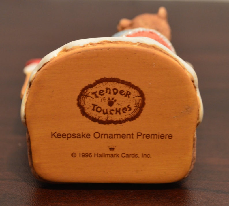 May include: A brown ceramic Christmas ornament featuring a bear wearing a red and white Santa hat and holding a red and white candy cane. The ornament is part of the "Tender Touches" collection and is a "Keepsake Ornament Premiere" from 1996 Hallmark Cards, Inc.