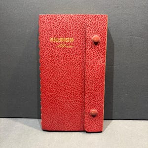 May include: A rectangular red View-Master Album with a textured, leather-like finish. The front cover features the gold-colored text "VIEW-MASTER Album" and includes a vertical flap secured by two circular red snap buttons on the right side.