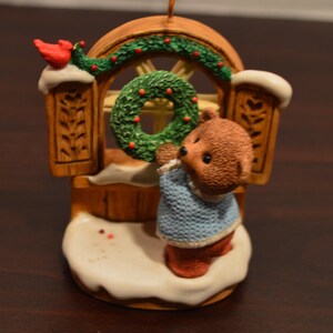 May include: A brown teddy bear wearing a blue sweater hangs a green wreath on a wooden door with a red cardinal perched on top. The ornament is decorated with white snow and red berries.