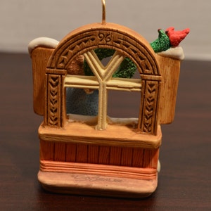 May include: A brown wooden window ornament with a white frame and a green wreath. The ornament has a red cardinal perched on top and the number "96" carved into the wood.