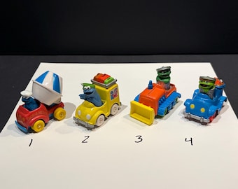 Puede incluir: Cuatro vehículos de juguete con personajes de Sesame Street, numerados del 1 al 4. El primero es una hormigonera roja con un personaje azul. El segundo es un coche amarillo con un personaje azul. El tercero es una excavadora naranja con un personaje verde. El cuarto es un coche azul con un personaje verde.
