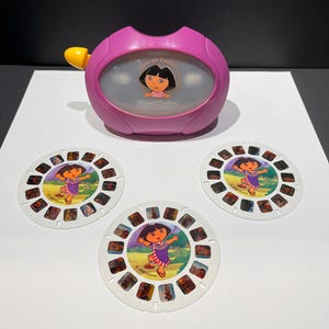 May include: A pink Dora the Explorer View-Master toy with a yellow advance lever and a translucent viewing screen featuring a Dora graphic. Included are three circular white image reels, each displaying multiple small color scenes of Dora.