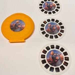 May include: A bright yellow plastic View-Master reel storage case and three circular white View-Master reels. Each reel features a central image of the animated character Remy from Ratatouille with the Eiffel Tower in the background.