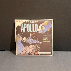 May include: A vintage Project Apollo View-Master box. The box features an illustration of an astronaut in space, a spacecraft, and the text "NASA's program for putting the first MAN on the MOON." The box also includes the text "STORY BOOKLET" and "21 VIEW MASTER Stereo Pictures."