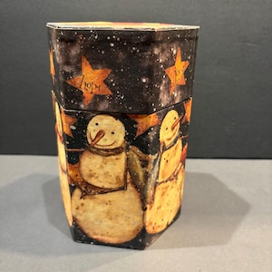 May include: A hexagonal decorative container featuring a dark night sky background with white speckles and orange stars labeled with the words hope and joy. The sides are decorated with illustrations of snowmen with orange carrot noses.