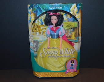 May include: A boxed Snow White doll with a yellow dress, blue bodice, and red cape. The box features a picture of Snow White and the Seven Dwarfs, and the text "Walt Disney's Snow White and the Seven Dwarfs".