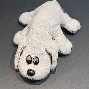 May include: A gray plush dog toy with black eyes and a black nose. The dog is lying down with its paws extended. A small red heart is embroidered on its back.