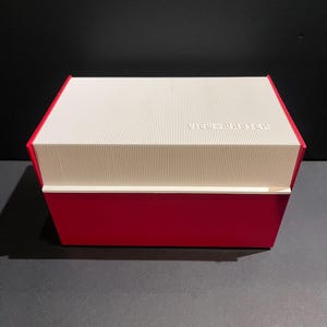 May include: A red rectangular box with a white, ridged lid. The lid has the words "VIEW-MASTER" printed on it. The box is sitting on a dark surface.