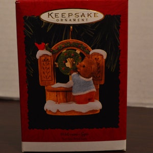 May include: A red and white box with a gold inscription that reads "Keepsake Ornament". The box features a picture of a brown bear wearing a blue sweater hanging a wreath on a wooden sign that reads "Welcome Sign".