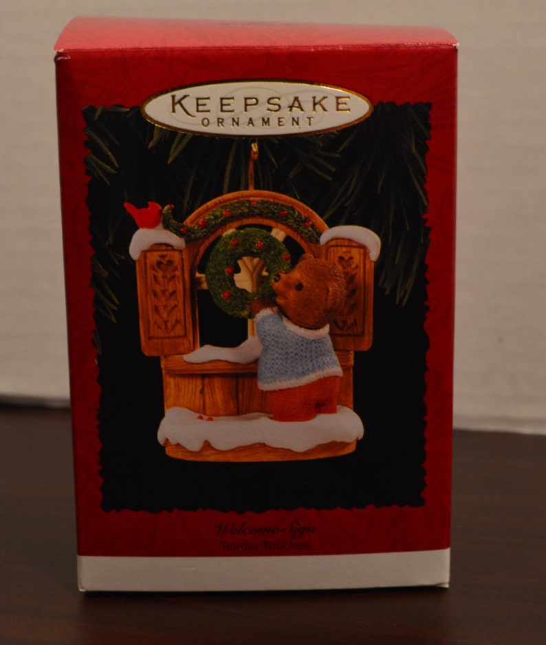 May include: A red and white box with a gold inscription that reads "Keepsake Ornament". The box features a picture of a brown bear wearing a blue sweater hanging a wreath on a wooden sign that reads "Welcome Sign".