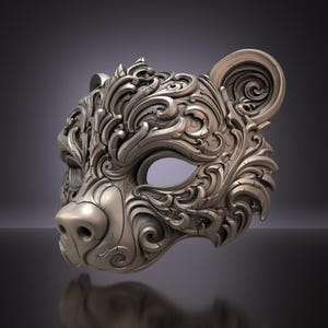 Może przedstawiać: Rzeźbiona maska niedźwiedzia z metalicznym wykończeniem w kolorze brązu, ozdobiona misternymi, wirującymi wzorami filigranowymi na całej powierzchni. Maska posiada wyraźny pysk, zaokrąglone uszy oraz jeden widoczny otwór na oko.