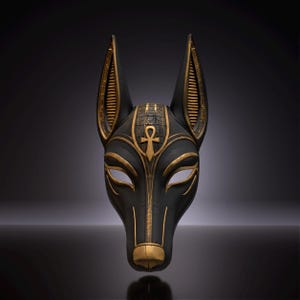 May include: A black and gold mask shaped like the head of a canine deity. The mask features pointed ears with ribbed detailing, an ankh symbol centered on the forehead, and intricate hieroglyphic-style carvings along the top and snout.