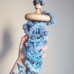 May include: A wooden drop spindle with a light blue and teal yarn wrap. The spindle has a rounded wooden whorl and a top with a small ball of yarn. The yarn is a mix of blues and purples.
