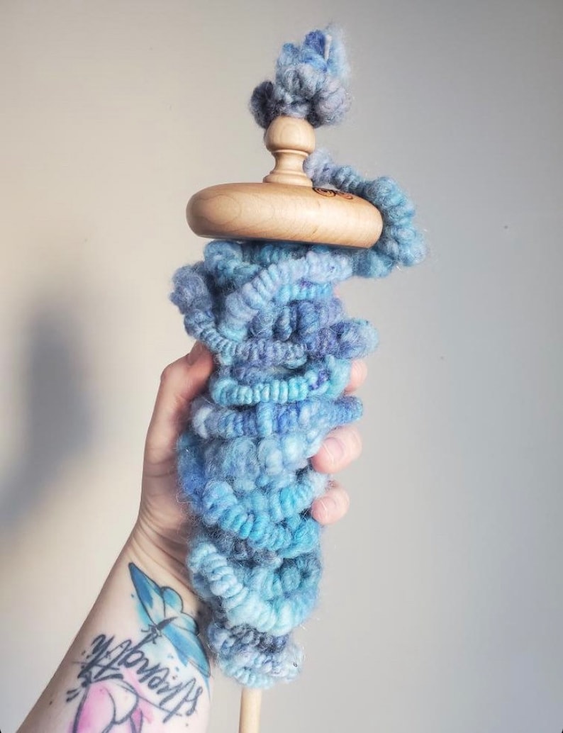 May include: A wooden drop spindle with a light blue and teal yarn wrap. The spindle has a rounded wooden whorl and a top with a small ball of yarn. The yarn is a mix of blues and purples.