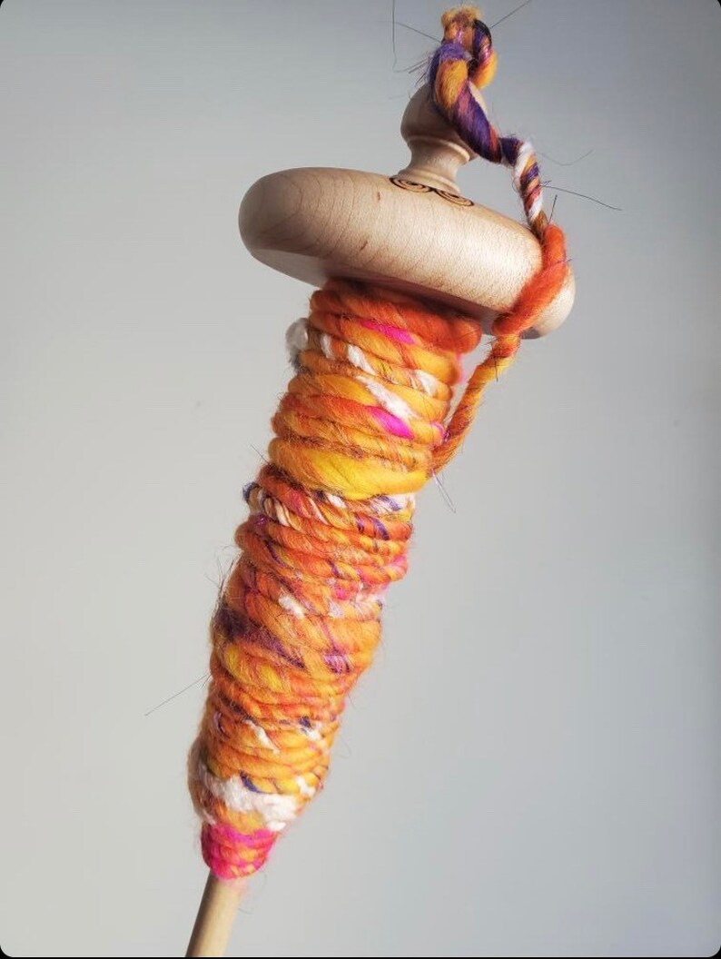 May include: A wooden drop spindle with a light-colored wooden whorl and shaft. The spindle is wrapped with vibrant orange, yellow, and pink hand-spun yarn. A twisted strand of colorful yarn is attached to the top.