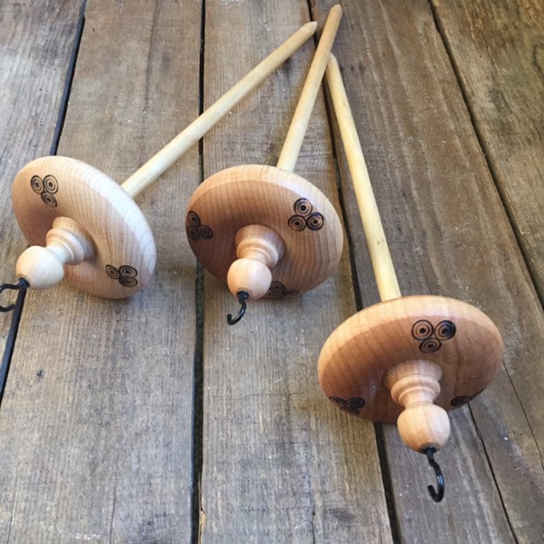 May include: Three wooden drop spindles with a natural finish. Each spindle features a round, light-colored wooden whorl with a decorative black design and a long, slender shaft. The spindles are arranged on a wooden surface.