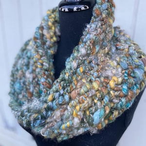 May include: A chunky, hand-knitted infinity scarf in a blend of teal, yellow, brown, and cream yarns. The scarf is displayed on a black mannequin head, showcasing its thick, textured weave. The color palette evokes a natural, earthy feel.
