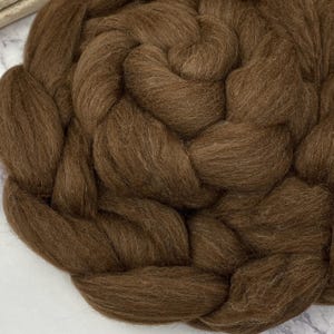May include: Close-up of a large, braided bundle of brown wool roving. The fibers are tightly packed and have a soft, textured appearance. The color is a rich, warm brown, suitable for various fiber art projects like felting or spinning.