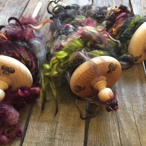 May include: Wooden drop spindles with a decorative design are shown with colorful, hand-dyed fiber. The fiber includes shades of purple, green, gray, and orange. The spindles are on a wooden surface.