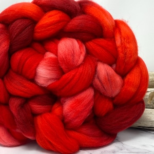 May include: Close-up of a large, braided bundle of vibrant red wool roving. The wool fibers are thick and soft, with variations in shades from deep crimson to bright scarlet. The texture is fluffy and inviting, perfect for crafting.