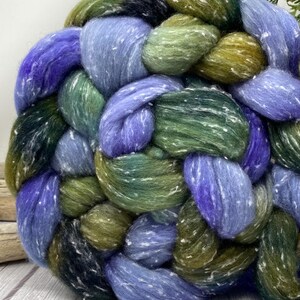 May include: Close-up of a braided yarn in shades of blue, green, and olive. The yarn has a speckled texture with white flecks throughout. The thick strands are interwoven, creating a textured, tactile appearance. The yarn is likely used for knitting or other fiber crafts.