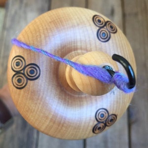 May include: A wooden drop spindle with a light wood finish. The spindle features three sets of black and blue circular designs. A strand of purple and blue yarn is wrapped around the spindle, with a black hook at the top.