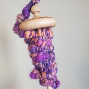 May include: A wooden drop spindle with a hand-spun yarn wrap. The yarn is a blend of purple, pink, and cream colors. The spindle has a smooth, rounded wooden whorl and a slender wooden shaft, perfect for fiber art projects.