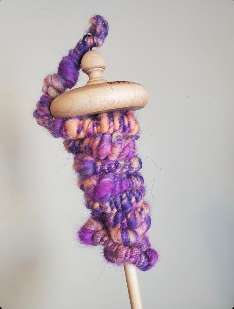 May include: A wooden drop spindle with a hand-spun yarn wrap. The yarn is a blend of purple, pink, and cream colors. The spindle has a smooth, rounded wooden whorl and a slender wooden shaft, perfect for fiber art projects.