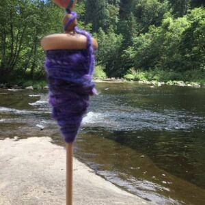 May include: A wooden drop spindle with a purple and blue yarn wrap. The spindle has a light brown wooden whorl and a long, thin wooden shaft. The yarn is a mix of dark and light purples and blues. The background is a river and trees.