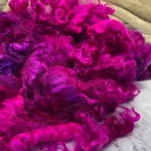 May include: Close-up of vibrant fuchsia-colored wool roving. The fiber is tightly curled, creating a textured appearance. The wool is a mix of bright pink and deep purple hues. The background includes a light-colored surface and driftwood.