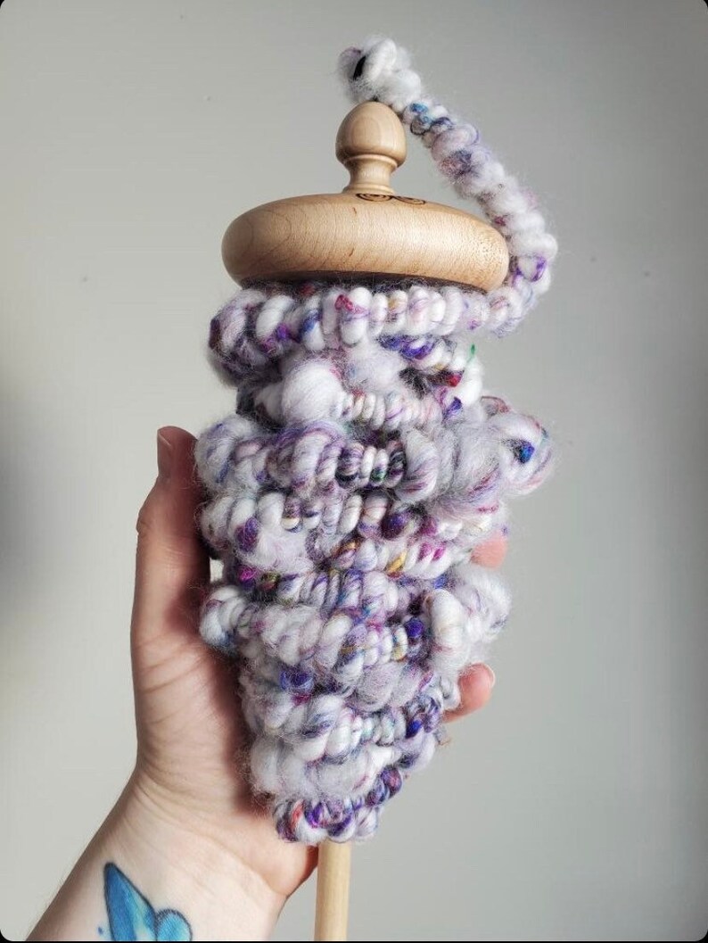 May include: A wooden drop spindle with a light-colored, rounded top and a long, thin wooden shaft. The spindle is wrapped with thick, textured yarn in shades of white, purple, and pink. The yarn is arranged in a conical shape, creating a visually appealing and functional tool.