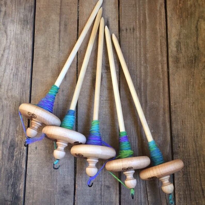 May include: Five wooden drop spindles with natural wood shafts and colorful yarn wraps in shades of blue, green, and purple. The spindles have a decorative design on the wooden base and a small hook at the bottom. The background is a rustic wooden surface.