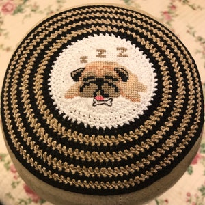 Puede incluir: Una kipá redonda tejida a ganchillo con anillos concéntricos alternos de hilo negro y beige. El centro muestra un parche circular blanco con un diseño de punto de cruz de un perro pug beige dormido con lengua rosa y un icono de hueso.