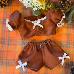 Puede incluir: Un conjunto de muñeca de dos piezas en color marrón que consta de una blusa de manga corta con ribete de encaje blanco, un lazo de cinta blanca central y pantalones cortos a juego con dos pequeños lazos blancos y un broche metálico.