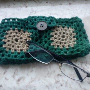 Può includere: Un astuccio per occhiali in crochet verde con un design in crochet beige e una chiusura a bottone marrone. Un paio di occhiali con montatura nera è appoggiato sopra l'astuccio.