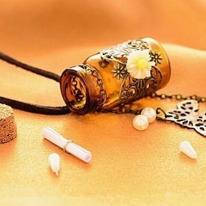 May include: A brown glass bottle with a metal floral design and a white flower charm hangs from a black cord. The bottle is attached to a metal butterfly charm and a white pearl bead. A cork stopper and a rolled up piece of paper are also included.